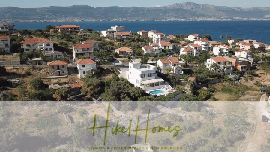 340m² Villa mit Pool auf der Insel Ciovo – vollständig möbiliert – auch Boutiquehotel möglich - Ein Küstendorf mit Häusern mit roten Dächern liegt an einem Hang und bietet Blick auf das blaue Meer und die Berge in der Ferne. Am unteren Bildrand sind die Texte „HikelHomes“ und „Traum- und Ferienimmobilien in Kroatien“ eingeblendet. (41626)