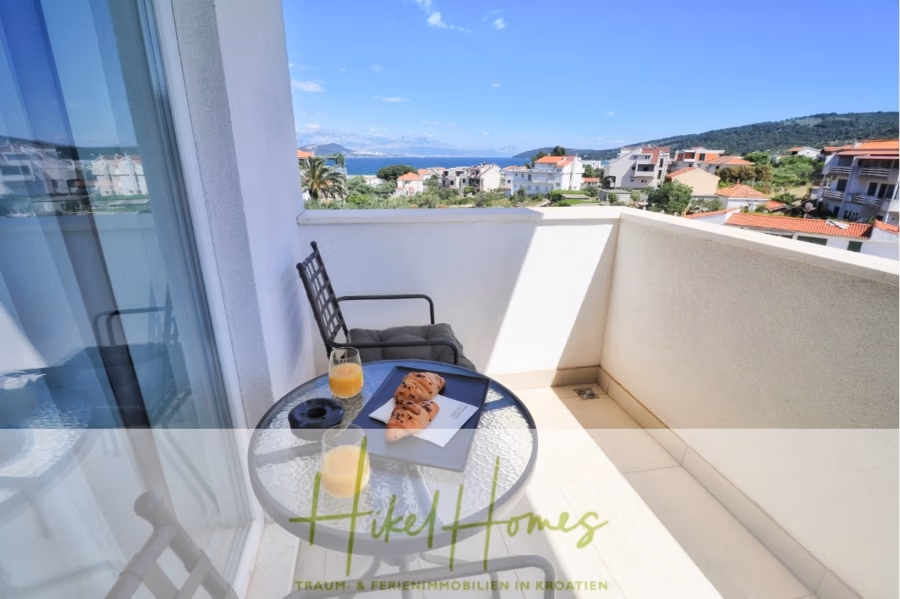 340m² Villa mit Pool auf der Insel Ciovo – vollständig möbiliert – auch Boutiquehotel möglich - Ein Balkon mit einem kleinen Tisch und zwei Stühlen bietet eine malerische Aussicht. Auf dem Tisch stehen zwei Gläser Orangensaft und Croissants auf einem Tablett. Im Hintergrund ist eine Küstenstadt mit Gebäuden, viel Grün und einem fernen Meerblick zu sehen. (41624)