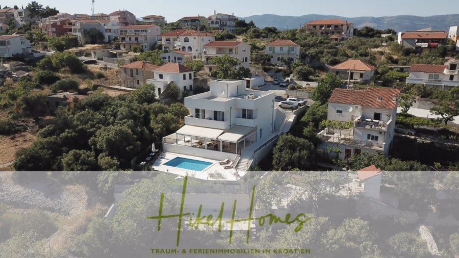 340m² Villa mit Pool auf der Insel Ciovo – vollständig möbiliert – auch Boutiquehotel möglich - Luftaufnahme eines Wohngebiets mit einem großen modernen Haus mit Pool im Vordergrund, umgeben von Grünflächen und mehreren weiteren Häusern mit roten Ziegeldächern. Im Hintergrund sind Hügel zu sehen. Textüberlagerung: „HKellHomes TRAUM- & FERIENIMMOBILIEN IN KROATIEN.“. (41633)