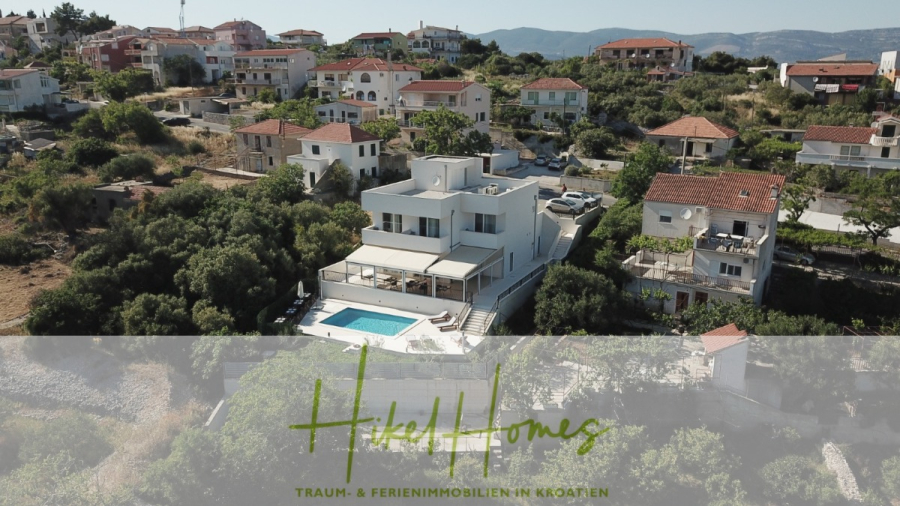 340m² Villa mit Pool auf der Insel Ciovo – vollständig möbiliert – auch Boutiquehotel möglich - Luftaufnahme einer modernen weißen Villa mit Swimmingpool, umgeben von Grünflächen und Nachbarhäusern in einem hügeligen Gebiet. Das Logo "HeleHomes" und der Text "TRAUM- & FERIENIMMOBILIEN IN KROATIEN" erscheinen am unteren Rand. (45542)