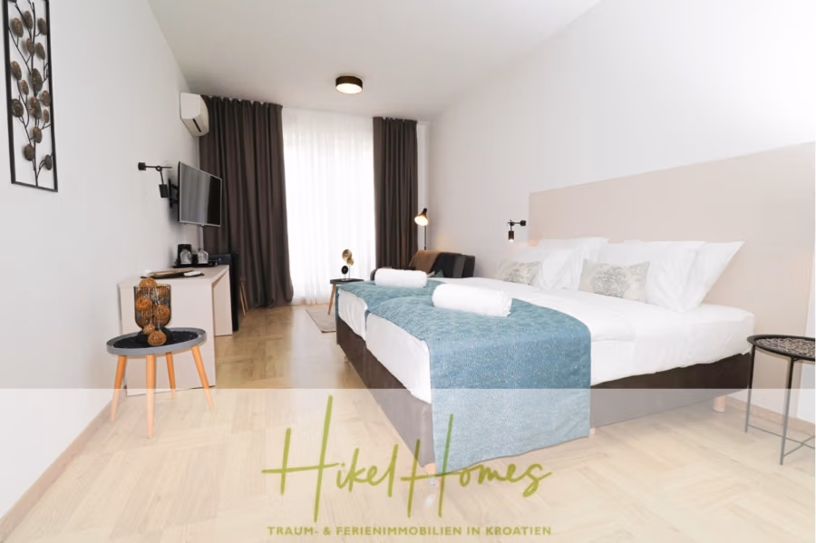 340m² Villa mit Pool auf der Insel Ciovo – vollständig möbiliert – auch Boutiquehotel möglich - Modernes Hotelzimmer mit großem Bett, weißer Bettwäsche und Kissen. Ein Flachbildfernseher hängt an der Wand, und ein kleiner Tisch mit Dekoartikeln steht bereit. Ein Fenster mit dunklen Vorhängen lässt Tageslicht herein. Textüberlagerung unten. (41630)