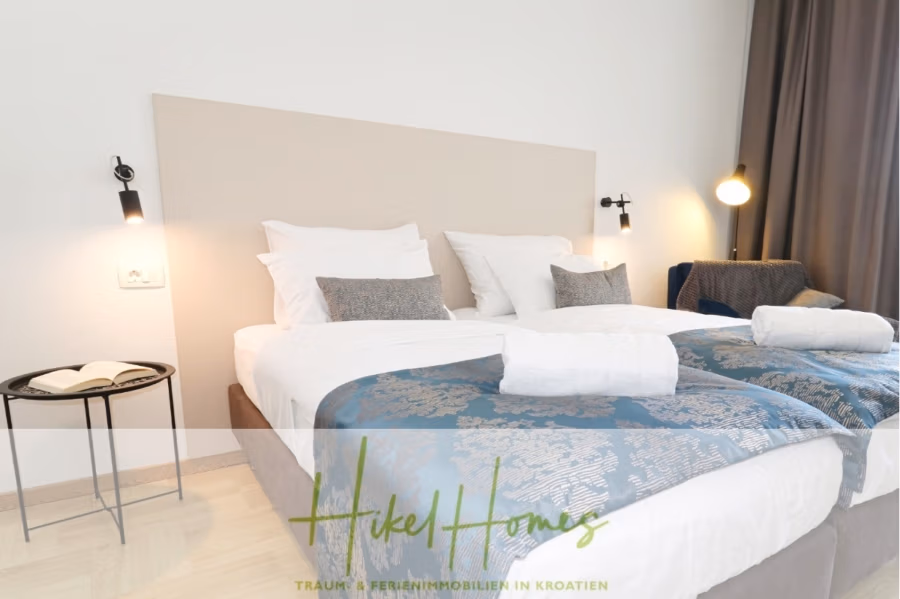 340m² Villa mit Pool auf der Insel Ciovo – vollständig möbiliert – auch Boutiquehotel möglich - Ein ordentlich gemachtes Hotelbett mit weißer Bettwäsche und blau gemusterten Überwürfen. Zwei Wandlampen flankieren das beigefarbene Kopfteil. Links steht ein kleiner runder Beistelltisch mit einem offenen Buch. Die Texteinblendung lautet „Hikel Homes, Traum- und Ferienimmobilien in Kroatien“. (41628)