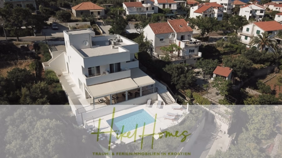 340m² Villa mit Pool auf der Insel Ciovo – vollständig möbiliert – auch Boutiquehotel möglich - Luftaufnahme eines modernen, weißen Hauses mit Pool in einem Wohngebiet mit viel Grün. Das Haus verfügt über mehrere Balkone und eine schattige Terrasse. Um das Anwesen herum befinden sich weitere Häuser mit roten Ziegeldächern. Text überlagert den unteren Bildrand. (41632)