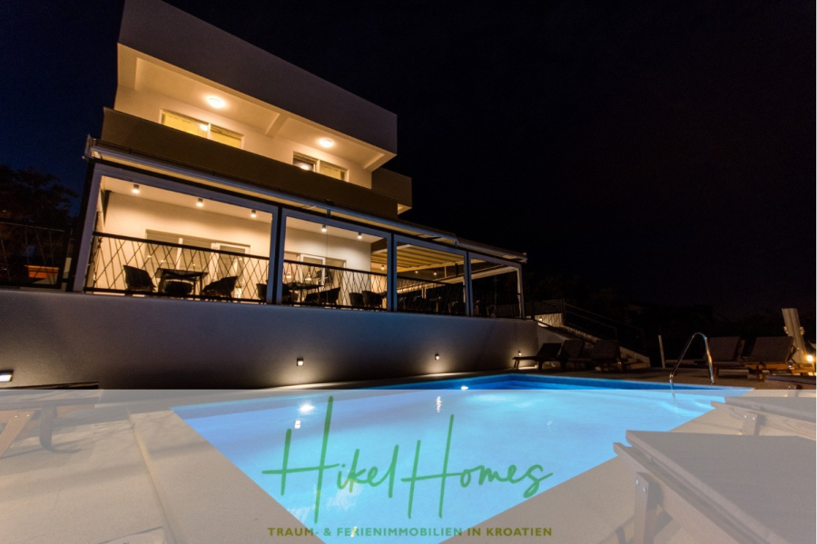 340m² Villa mit Pool auf der Insel Ciovo – vollständig möbiliert – auch Boutiquehotel möglich - Eine moderne, gut beleuchtete Villa bei Nacht mit großen Balkonen und einem leuchtenden Außenschwimmbad im Vordergrund. Der Text "Hikel Homes - Traum & Ferienimmobilien in Kroatien" überlagert den unteren Teil des Bildes. (45549)