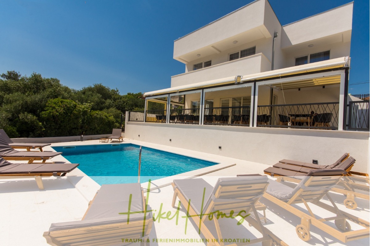 Moderne weiße Villa mit einer großen überdachten Terrasse, Liegestühlen am Pool und einem Swimmingpool, umgeben von Grün unter einem klaren blauen Himmel. Textüberlagerung: "Heike Homes" in Grün und Gelb. - 340m² Villa mit Pool auf der Insel Ciovo – vollständig möbiliert – auch Boutiquehotel möglich