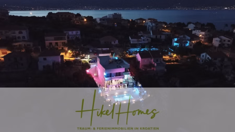 340m² Villa mit Pool auf der Insel Ciovo – vollständig möbiliert – auch Boutiquehotel möglich - Eine nächtliche Luftaufnahme eines Wohngebiets am Meer. Ein zentrales Haus ist in verschiedenen Farben hell erleuchtet, mit einem leuchtenden Pool. Die umliegenden Häuser sind schwach beleuchtet. Die Szene fängt eine ruhige Küstenatmosphäre ein. Der Textüberzug lautet „Hikel Homes“. (41641)