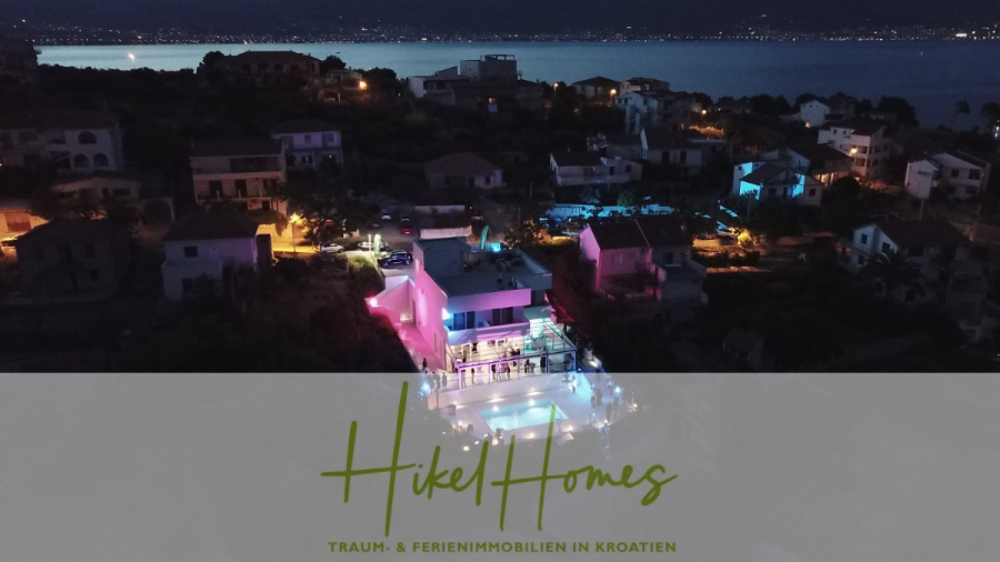 340m² Villa mit Pool auf der Insel Ciovo – vollständig möbiliert – auch Boutiquehotel möglich - Nächtliche Luftaufnahme einer modernen Villa mit einem von bunten Lichtern beleuchteten Pool, umgeben von anderen Häusern in der Nähe einer Küste in Kroatien. Der überlagerte Text lautet: "Hikel Homes - Traum- & Ferienimmobilien in Kroatien. (45553)