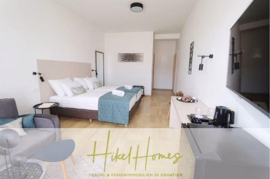 340m² Villa mit Pool auf der Insel Ciovo – vollständig möbiliert – auch Boutiquehotel möglich - Modernes Hotelzimmer mit großem Bett, dekorativen Kissen und türkisfarbenen Akzenten. Eine kleine Sitzecke mit grauem Stuhl und rundem Tisch ist sichtbar. Das Zimmer verfügt über einen wandmontierten Fernseher und einen Wasserkocher auf einem Schrank. Das Bild enthält unten ein Logo und deutschen Text. (41627)