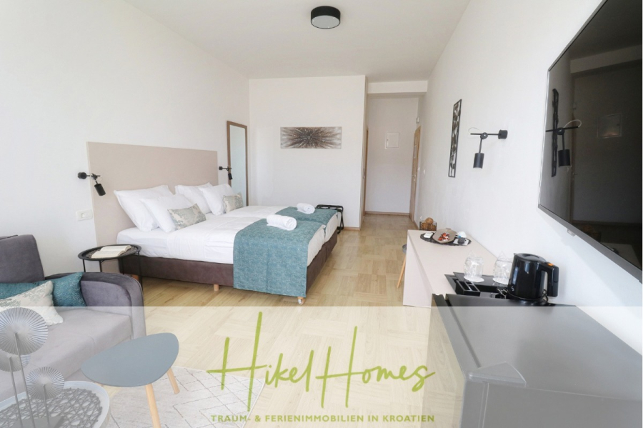 340m² Villa mit Pool auf der Insel Ciovo – vollständig möbiliert – auch Boutiquehotel möglich - Ein helles, modernes Hotelzimmer mit zwei Betten, weißer Bettwäsche, blaugrünen Decken, einem an der Wand montierten Fernseher, einem Sessel, einer Kaffeestation und hellen Holzböden. Im Hintergrund sind ein Flur und Wandkunst zu sehen. (45534)