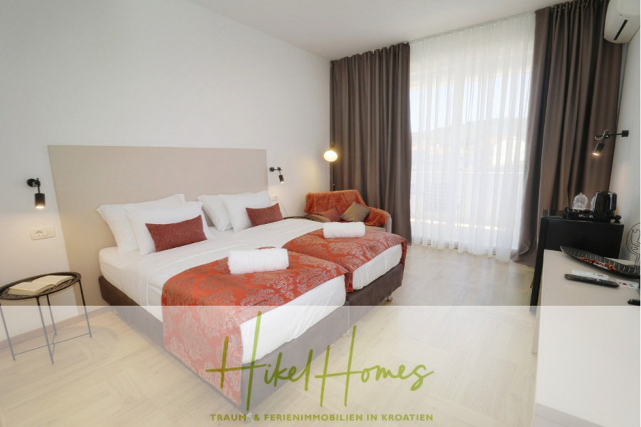 340m² Villa mit Pool auf der Insel Ciovo – vollständig möbiliert – auch Boutiquehotel möglich - Ein modernes Hotelzimmer mit zwei aneinandergeschobenen Einzelbetten, roter und weißer Bettwäsche, einem kleinen Sofa, Nachttischen, einer Wandlampe, einem großen Fenster mit Vorhängen und einem Schreibtisch mit einer Kaffeestation. Der Schriftzug "Hikel Homes" ist überlagert. (45545)