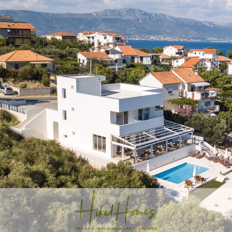 340m² Villa mit Pool auf der Insel Ciovo – vollständig möbiliert – auch Boutiquehotel möglich - Moderne weiße Villa mit verglaster Terrasse und Pool, umgeben von viel Grün. Die Villa liegt in einem Wohngebiet mit anderen Häusern in der Nähe und bietet Blick auf eine ferne Bergkette und das Meer unter einem klaren blauen Himmel. (41623)