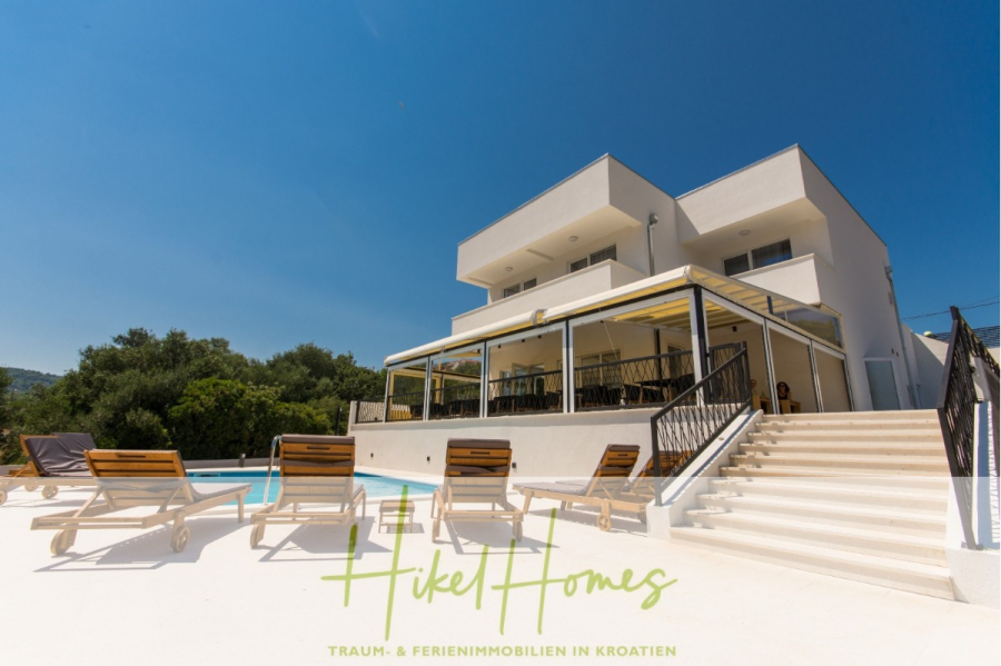 340m² Villa mit Pool auf der Insel Ciovo – vollständig möbiliert – auch Boutiquehotel möglich - Moderne weiße Villa mit großen Balkonen, Liegestühlen am Pool und einem Swimmingpool unter blauem Himmel. Umgeben von Grünflächen. Der Text am unteren Rand lautet: "Hikel Homes TRAUM- & FERIENIMMOBILIEN IN KROATIEN.". (45528)