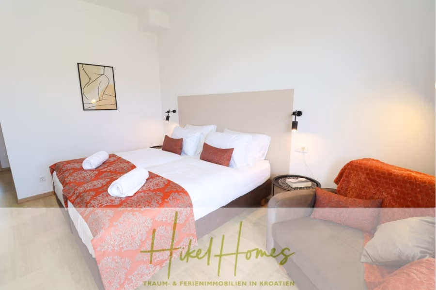 340m² Villa mit Pool auf der Insel Ciovo – vollständig möbiliert – auch Boutiquehotel möglich - Ein gemütliches Hotelzimmer mit einem großen Bett, ausgestattet mit rot-weißer Bettwäsche und passenden Kissen. Daneben steht ein Sessel, bedeckt mit einer roten Decke. Minimalistische Kunstwerke schmücken die weiße Wand, beleuchtet von zwei Wandlampen. (41637)