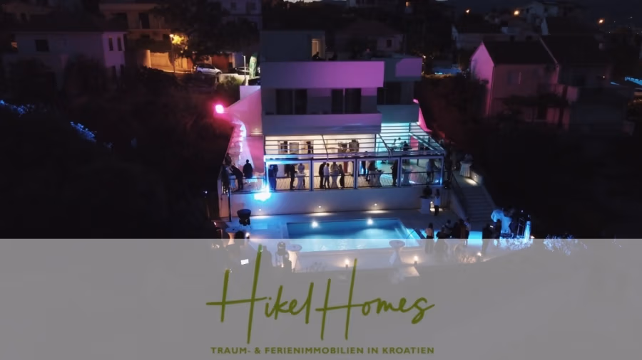 340m² Villa mit Pool auf der Insel Ciovo – vollständig möbiliert – auch Boutiquehotel möglich - Ein modernes Haus mit großen Glasfenstern ist nachts beleuchtet und von Bäumen umgeben. Menschen stehen auf einem Balkon mit Blick auf einen beleuchteten Pool. Das Bild enthält den Text „Hikel Homes – Traum- & Ferienimmobilien in Kroatien“. (41642)