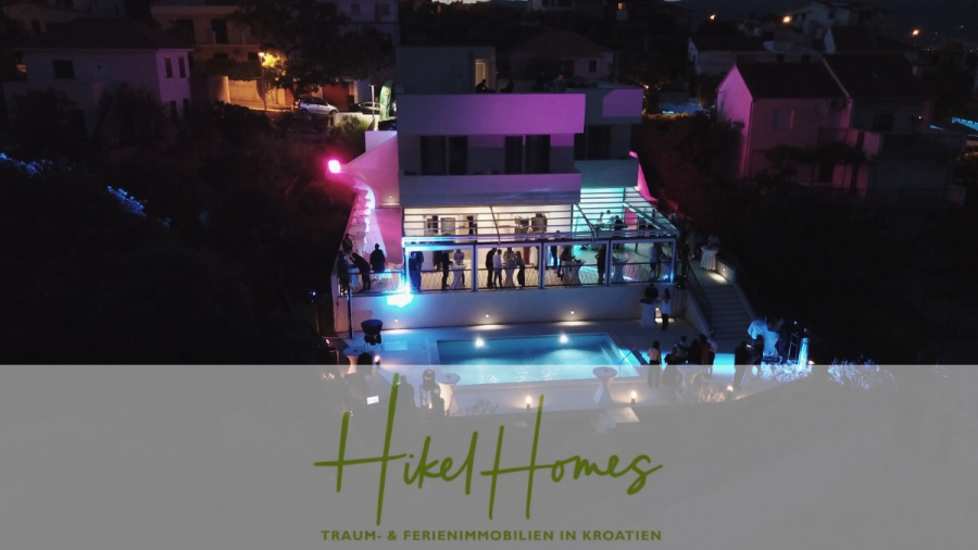 340m² Villa mit Pool auf der Insel Ciovo – vollständig möbiliert – auch Boutiquehotel möglich - Ein modernes Haus in Kroatien, das nachts mit violetten und blauen Lichtern beleuchtet ist, mit Menschen, die sich auf einem Balkon und um einen Pool versammelt haben. Der Text "Hikel Homes: Traum- & Ferienimmobilien in Kroatien" erscheint am unteren Rand. (45554)