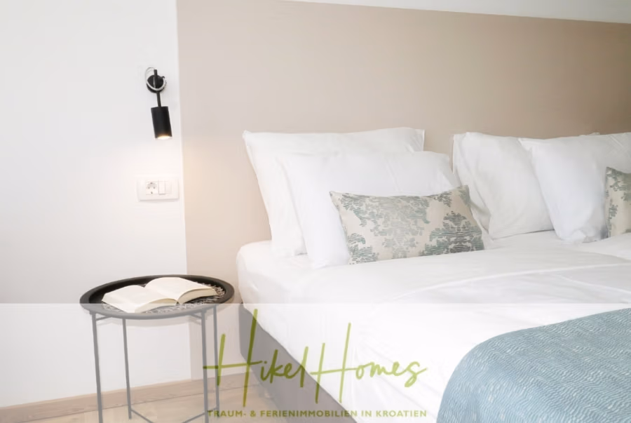 340m² Villa mit Pool auf der Insel Ciovo – vollständig möbiliert – auch Boutiquehotel möglich - Ein modernes Schlafzimmer mit einem ordentlich gemachten Bett und weißen, gemusterten Kissen. Daneben steht ein kleiner runder Tisch mit einem offenen Buch und einer Wandleuchte. Das Zimmer ist minimalistisch und in neutralen Farben gestaltet. (41631)