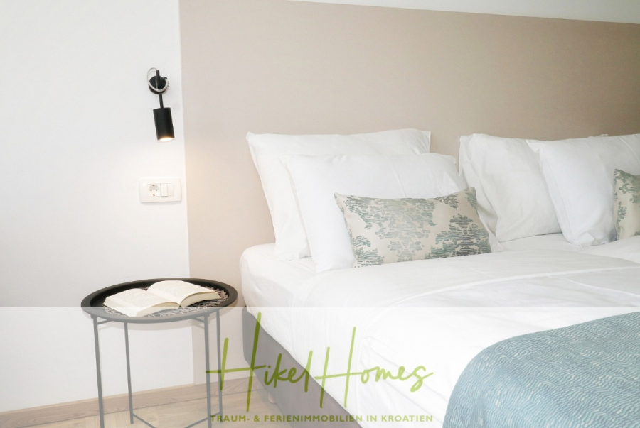 340m² Villa mit Pool auf der Insel Ciovo – vollständig möbiliert – auch Boutiquehotel möglich - Ein ordentlich gemachtes Bett mit weißer Bettwäsche und gemusterten Kissen neben einem runden schwarzen Beistelltisch, auf dem ein aufgeschlagenes Buch liegt, unter einer an der Wand befestigten Leselampe in einem hellen, modernen Schlafzimmer. (45540)