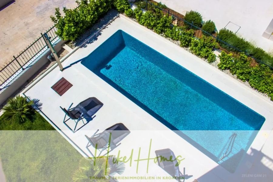 Villa im Mittelmeerstil mit 3 SZ, Pool, Dachterrasse mit Meerblick (1) - Ein moderner, rechteckiger Swimmingpool mit klarem, blauem Wasser, umgeben von einem weißen Deck. In der Nähe befinden sich zwei Liegestühle mit Beistelltischen, und der Bereich ist von Grün umgeben. Ein Logo ist teilweise am Boden sichtbar. (45816)