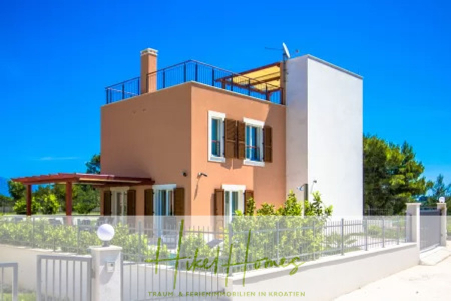 Villa im Mittelmeerstil mit 3 SZ, Pool, Dachterrasse mit Meerblick (1) - Ein modernes, zweistöckiges Haus mit braunen und weißen Außenwänden, hölzernen Fensterläden, einer Dachterrasse und einem eingezäunten Hof, vor einem strahlend blauen Himmel und umgeben von Grün. (45821)