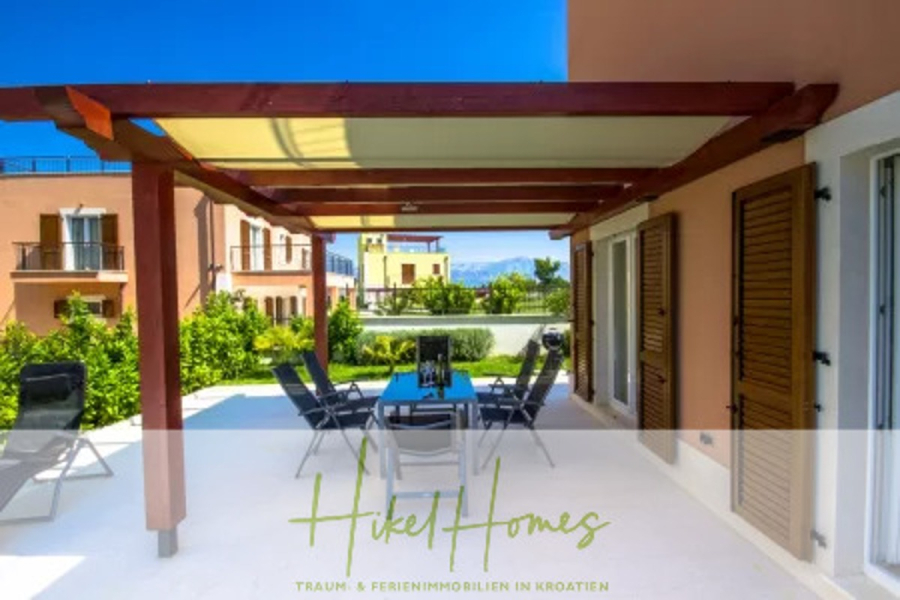 Villa im Mittelmeerstil mit 3 SZ, Pool, Dachterrasse mit Meerblick (1) - Eine geräumige Außenterrasse mit einem Esstisch und Stühlen unter einer hölzernen Pergola, die an ein modernes Haus mit braunen Fensterläden, üppigem Grün und anderen Häusern im Hintergrund angebaut ist. Der Text lautet "Hikel Homes. (45818)