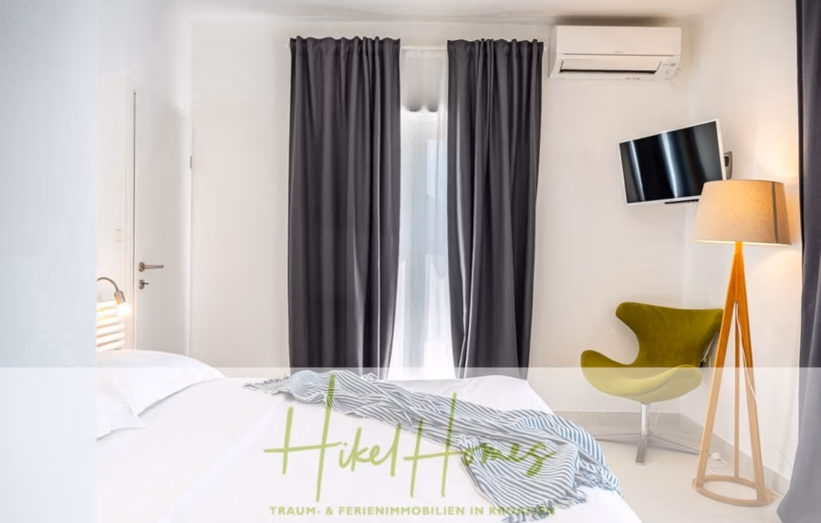 2 Meerblick Villen + freies Grundstück ideal zur Vermietungauf der Insel Dugi Otok jeweils mit Pool - Ein modernes, helles Schlafzimmer mit einem großen Bett, dunkelgrauen Vorhängen, einem an der Wand montierten Flachbildfernseher und einem grünen Stuhl. Neben dem Fernseher steht eine hohe Lampe. Das Zimmer hat weiße Wände und eine Klimaanlage. (42897)