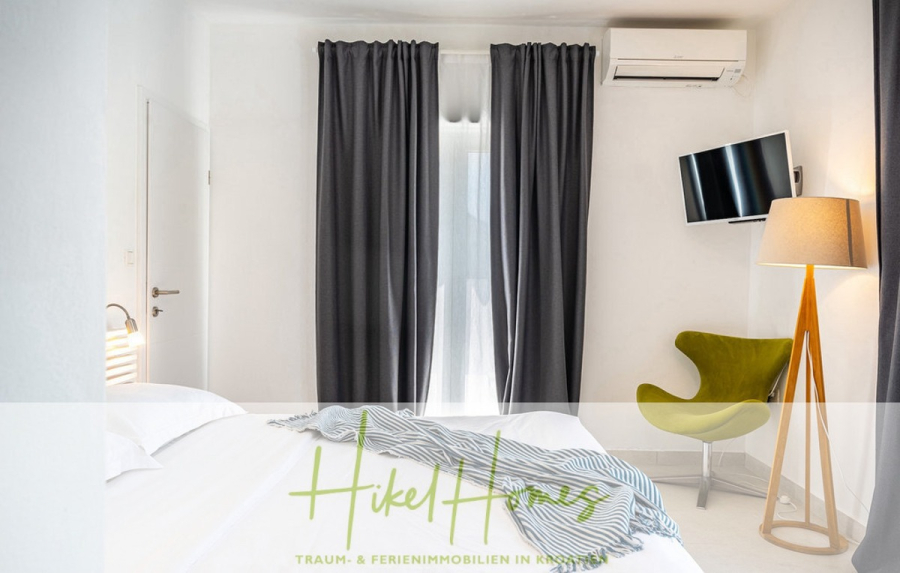 2 Meerblick Villen + freies Grundstück ideal zur Vermietungauf der Insel Dugi Otok jeweils mit Pool - Minimalistisches Schlafzimmer mit einem weißen Bett, grauen Vorhängen, einer Stehlampe, einem grünen Stuhl, einem an der Wand montierten Fernseher und einer Klimaanlage. Sanftes natürliches Licht fällt durch ein Fenster hinter den Vorhängen ein. (44741)