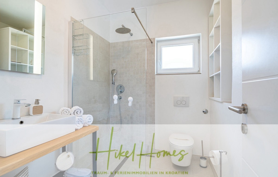 2 Meerblick Villen + freies Grundstück ideal zur Vermietungauf der Insel Dugi Otok jeweils mit Pool - Modernes Badezimmer mit Glasdusche, wandmontierter Toilette, weißem Waschbecken auf einer hölzernen Arbeitsplatte, Handtüchern und einem kleinen Fenster. Der Raum ist hell und minimalistisch eingerichtet. Das Logo "Hikel Homes" überlagert den unteren Teil des Bildes. (44739)