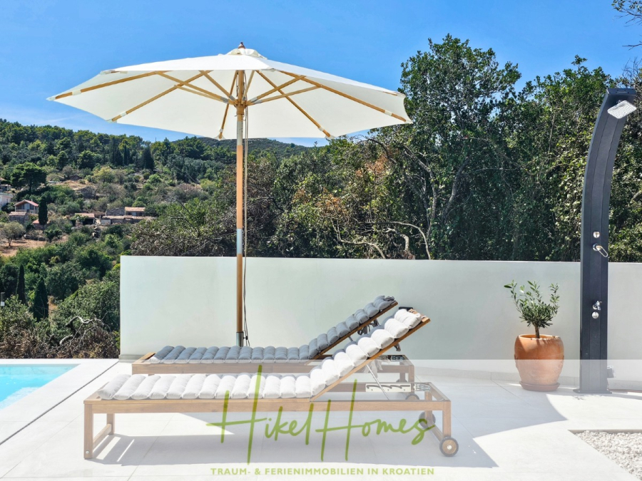 2 Meerblick Villen + freies Grundstück ideal zur Vermietungauf der Insel Dugi Otok jeweils mit Pool - Eine Szene am Pool mit zwei Liegestühlen unter einem großen weißen Sonnenschirm, einer Topfpflanze, einer Außendusche und einer malerischen Aussicht auf Hügel und Bäume. Der Text am unteren Rand lautet "Hikel Homes" und "TRAUM- & FERIENIMMOBILIEN IN KROATIEN". (44749)