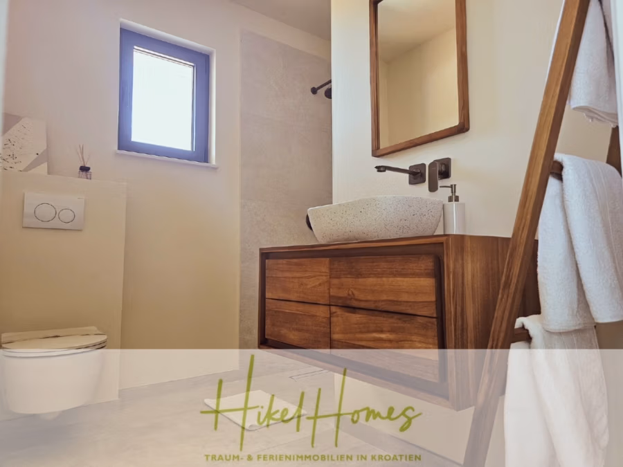 2 Meerblick Villen + freies Grundstück ideal zur Vermietungauf der Insel Dugi Otok jeweils mit Pool - Modernes Badezimmer mit Holzwaschtisch und Steinbecken, Wandarmatur, Handtuchleiter mit weißen Handtüchern und kleinem Fenster. Unten befindet sich das Logo „Hikel Homes – Traum- & Ferienimmobilien in Kroatien“. (42901)