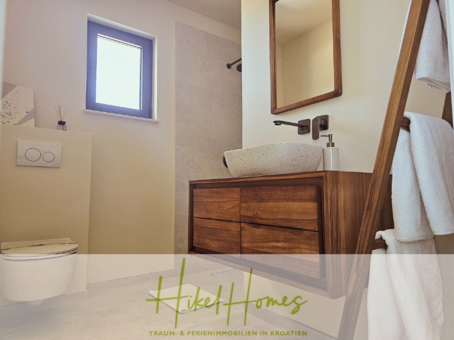 2 Meerblick Villen + freies Grundstück ideal zur Vermietungauf der Insel Dugi Otok jeweils mit Pool - Modernes Badezimmer mit einem hölzernen Waschtisch, einem Gefäßwaschbecken, einem Wandwasserhahn, einer Handtuchleiter und einem kleinen Fenster. Der Text "Hikel Homes Traum- & Ferienimmobilien in Kroatien" erscheint am unteren Rand. (44745)