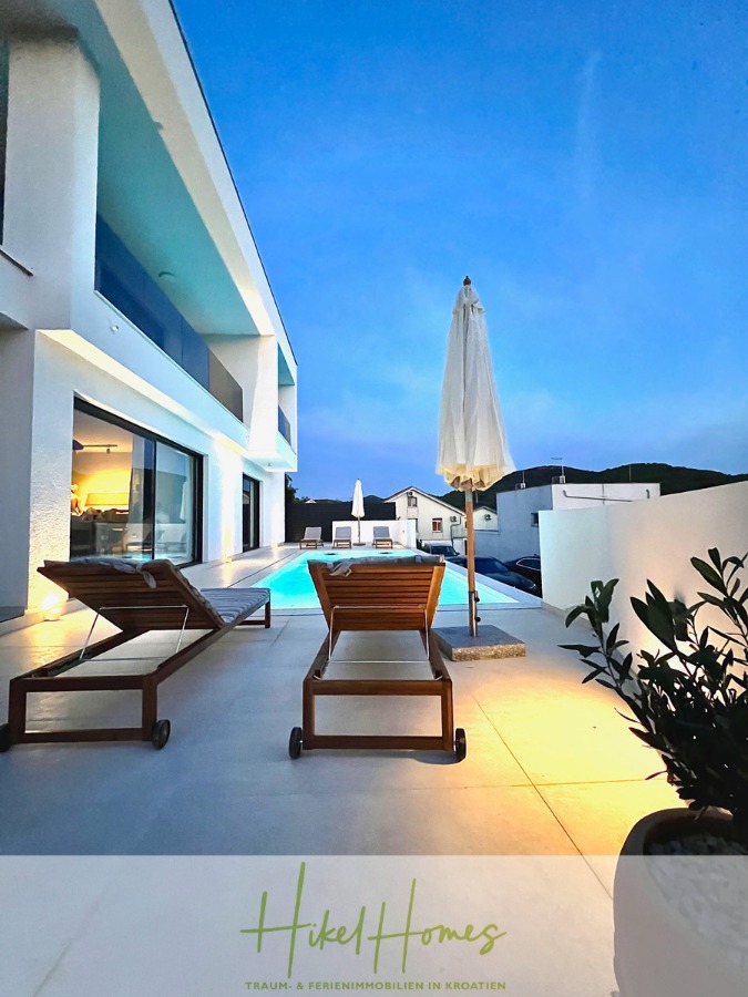 2 Meerblick Villen + freies Grundstück ideal zur Vermietungauf der Insel Dugi Otok jeweils mit Pool - Moderne Außenterrasse mit zwei hölzernen Liegestühlen neben einem Pool, beleuchtet von Abendlichtern. Ein geschlossener Sonnenschirm steht zwischen den Stühlen. Im Hintergrund sind moderne weiße Gebäude und Hügel zu sehen. (44759)
