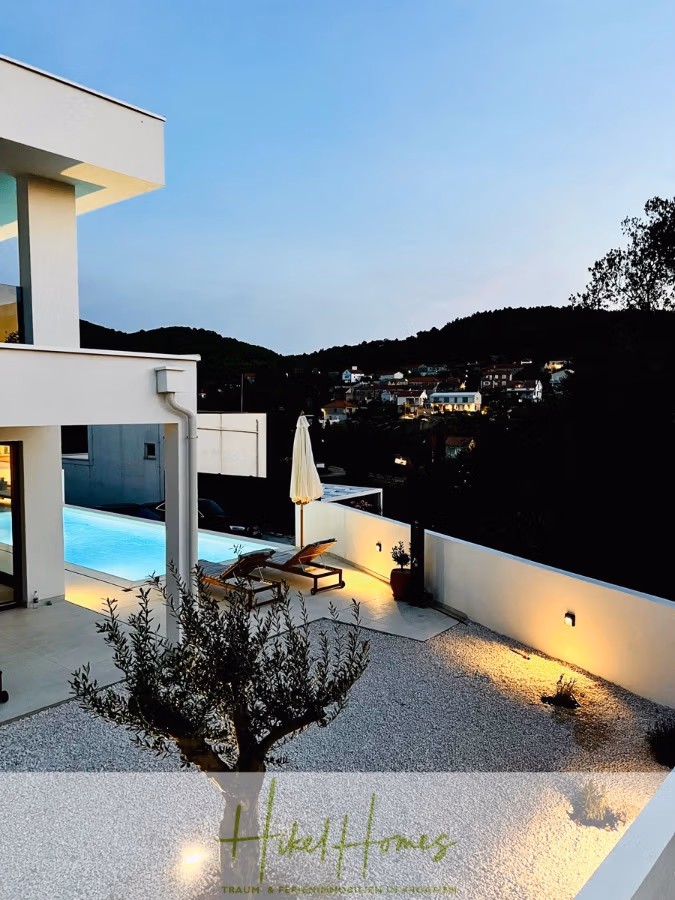 2 Meerblick Villen + freies Grundstück ideal zur Vermietungauf der Insel Dugi Otok jeweils mit Pool - Modernes Haus mit elegantem Design, einem leuchtenden Swimmingpool und Sitzgelegenheiten im Freien. Im Vordergrund steht ein kleiner Baum, umgeben von Kies. Hügel und ferne Stadtlichter sind unter einem Abendhimmel zu sehen. (42912)