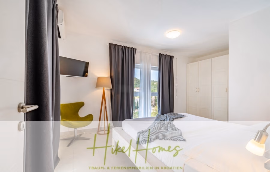 2 Meerblick Villen + freies Grundstück ideal zur Vermietungauf der Insel Dugi Otok jeweils mit Pool - Helles, modernes Schlafzimmer mit grünem Stuhl, Wandfernseher, großem Kleiderschrank und einem Fenster mit transparenten, dunklen Vorhängen. Das Bett ist ordentlich gemacht, und in der Ecke steht eine Stehlampe. Der Blick nach draußen ist grün. Unten ist das Hikel Homes-Logo zu sehen. (42895)