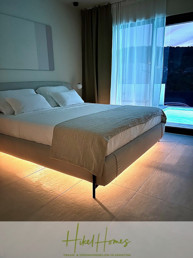 2 Meerblick Villen + freies Grundstück ideal zur Vermietungauf der Insel Dugi Otok jeweils mit Pool - Ein modernes Schlafzimmer mit einem großen Bett mit weicher Bettwäsche und Unterbettbeleuchtung, neben einem großen Fenster mit durchsichtigen Vorhängen, das den Blick auf die Hügel und den Pool draußen freigibt. Das Logo von "Hikel Homes" erscheint am unteren Rand. (44756)