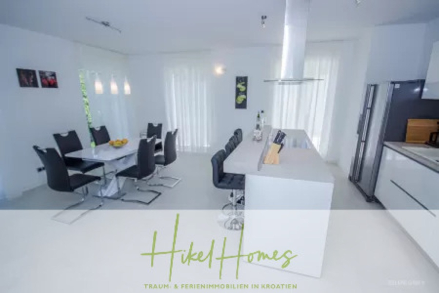 Villa im Mittelmeerstil mit 3 SZ, Pool, Dachterrasse mit Meerblick (7) - Moderne Küche und Essbereich mit weißer Insel und Barhockern, schwarzen Esszimmerstühlen um einen Tisch, großen Fenstern mit durchsichtigen Vorhängen und schlanken Geräten. Die Textüberlagerung lautet "Hikel Homes". (44836)