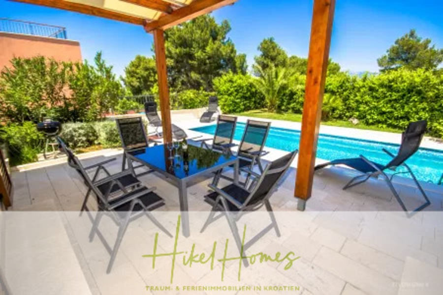 Villa im Mittelmeerstil mit 3 SZ, Pool, Dachterrasse mit Meerblick (7) - Terrasse im Freien mit einem Esstisch aus Glas und schwarzen Stühlen unter einer hölzernen Pergola neben einem Swimmingpool, umgeben von üppigem Grün und strahlend blauem Himmel. Die Textüberlagerung unten lautet "Hikel Homes" mit zusätzlichem deutschen Text. (44827)