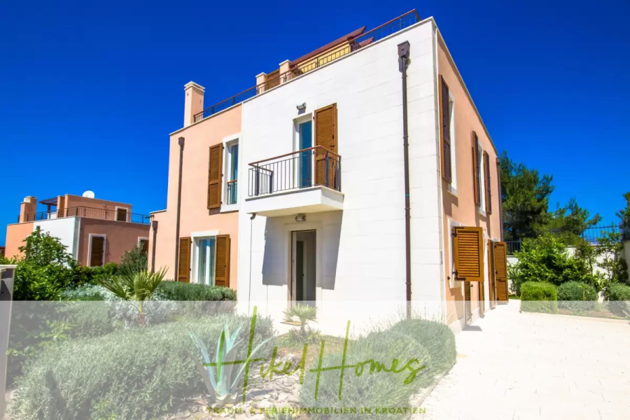 Villa im Mittelmeerstil mit 3 SZ, Pool, Dachterrasse mit Meerblick (7) - Modernes zweistöckiges Haus mit creme- und pfirsichfarbenem Äußeren, braunen Fensterläden, kleinen Balkonen und umgeben von grünen Sträuchern und Pflanzen unter einem klaren blauen Himmel. Das Logo "Vila Homes" ist am unteren Rand eingeblendet. (44825)