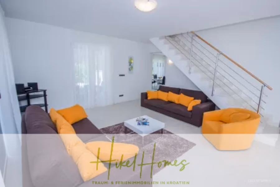 Villa im Mittelmeerstil mit 3 SZ, Pool, Dachterrasse mit Meerblick (7) - Modernes Wohnzimmer mit zwei grauen Sofas, einem orangefarbenen Sessel und orangefarbenen Kissen. Ein kleiner weißer Couchtisch steht auf einem grauen Teppich. Eine Treppe mit Metallgeländer führt ins Obergeschoss. Helles Tageslicht fällt durch transparente Vorhänge. (41661)