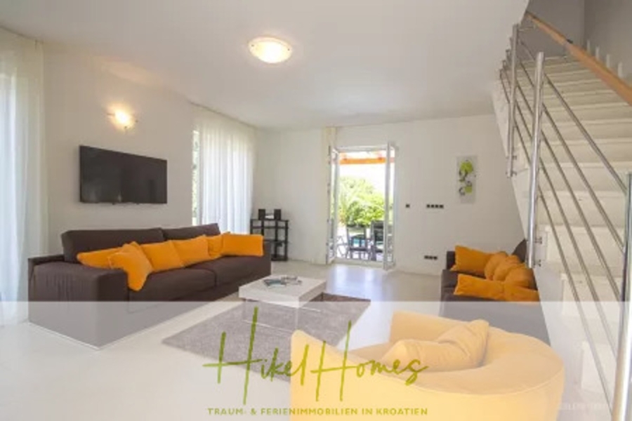 Villa im Mittelmeerstil mit 3 SZ, Pool, Dachterrasse mit Meerblick (7) - Modernes Wohnzimmer mit weißen Wänden und großen Fenstern. Es verfügt über ein braunes Sofa mit gelben Kissen, einen runden gelben Sessel, einen Wandfernseher und eine Treppe. Eine Glasschiebetür führt auf eine begrünte Terrasse. Der Text lautet „Hike Homes“. (41660)