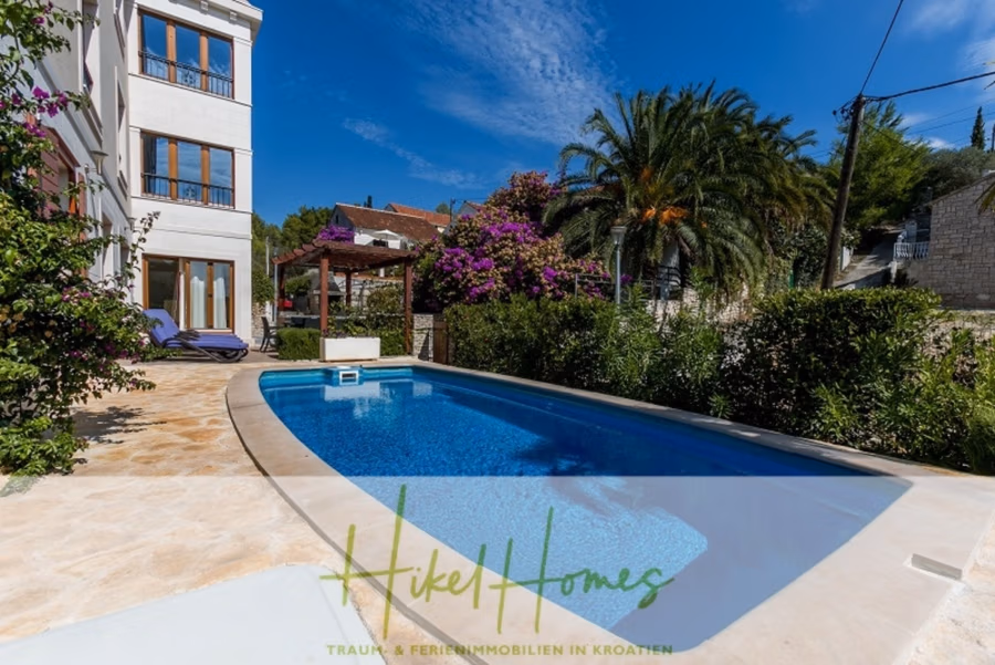 Villa mit 3 großzügigen Appartements mit jeweils 3 SZ, Küche, 2 x Pool, … - Eine ruhige Außenszene zeigt einen rechteckigen Pool neben einem weißen Gebäude mit großen Fenstern. Palmen und leuchtend violette Blumen bereichern die Atmosphäre. Im Hintergrund befindet sich eine Pergola mit Sitzgelegenheiten unter einem strahlend blauen Himmel. (41134)
