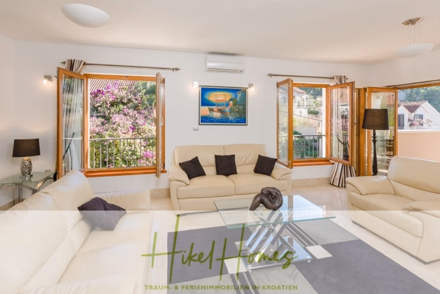 Villa mit 3 großzügigen Appartements mit jeweils 3 SZ, Küche, 2 x Pool, … - Helles Wohnzimmer mit weißen Sofas, Glascouchtisch und großen Fenstern mit offenen Holzläden. Kunst an der Wand und dekorative Pflanzen im Außenbereich sorgen für tropisches Flair. Der Text unten lautet: „Hikel Homes – Traum- & Ferienimmobilien in Kroatien.“. (41142)