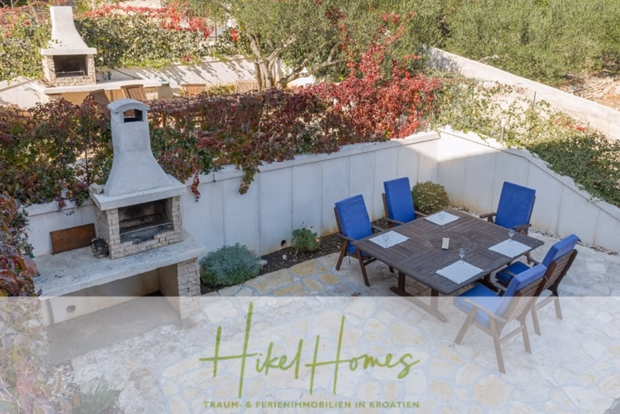Villa mit 3 großzügigen Appartements mit jeweils 3 SZ, Küche, 2 x Pool, … - Eine Terrasse mit einem dunklen Holztisch und sechs blauen Polsterstühlen auf Steinboden. In der Ecke ist ein eingebauter Steingrill zu sehen. Der Bereich ist von üppigem Grün und Rot umgeben. Der Text lautet: „Hikel Homes Traum- & Ferienimmobilien in Kroatien.“. (41138)