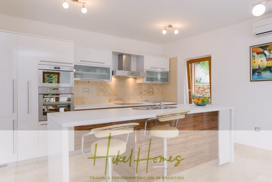 Villa mit 3 großzügigen Appartements mit jeweils 3 SZ, Küche, 2 x Pool, … - Moderne Küche mit weißen Schränken und Geräten, beiger Küchenrückwand und einer Kücheninsel aus Holz. Zwei beige Barhocker stehen auf der Insel. Ein Fenster bietet Blick auf den Garten, an der Wand hängt ein farbenfrohes Gemälde. Deckenleuchten betonen den hellen Raum. (41136)