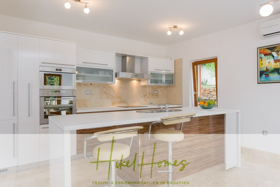 Villa mit 3 großzügigen Appartements mit jeweils 3 SZ, Küche, 2 x Pool, … - Moderne Küche mit weißen Schränken, Einbaugeräten, einer holzverkleideten Insel mit zwei beigen Barhockern, einer Obstschale und einem Fenster. Die Wände sind mit Kunstwerken dekoriert und die warme Beleuchtung sorgt für eine gemütliche Atmosphäre. (46266)