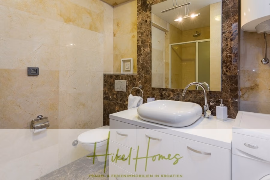 Villa mit 3 großzügigen Appartements mit jeweils 3 SZ, Küche, 2 x Pool, … - Modernes Badezimmer mit Wänden aus beige-braunem Marmor. Eine weiße Waschtischplatte trägt ein elegantes, ovales Waschbecken und einen glänzenden Wasserhahn. Im Hintergrund befindet sich eine Glasduschkabine, und eine Toilette mit wandmontiertem Spülsystem. Stilvolles und klares Design. (41141)