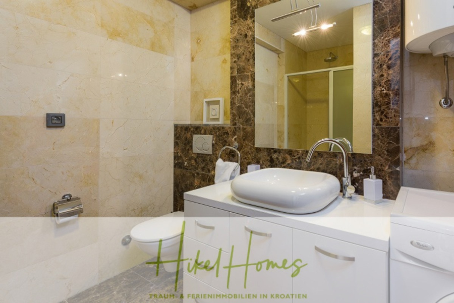 Villa mit 3 großzügigen Appartements mit jeweils 3 SZ, Küche, 2 x Pool, … - Modernes Badezimmer mit beigen und braunen Marmorwänden, einem weißen Waschbecken mit einem eleganten Wasserhahn auf einem Schrank, einer Toilette, einer Glasduschabtrennung und Toilettenartikeln auf dem Tresen. Das Logo von "Hikel Homes" erscheint am unteren Rand. (46270)