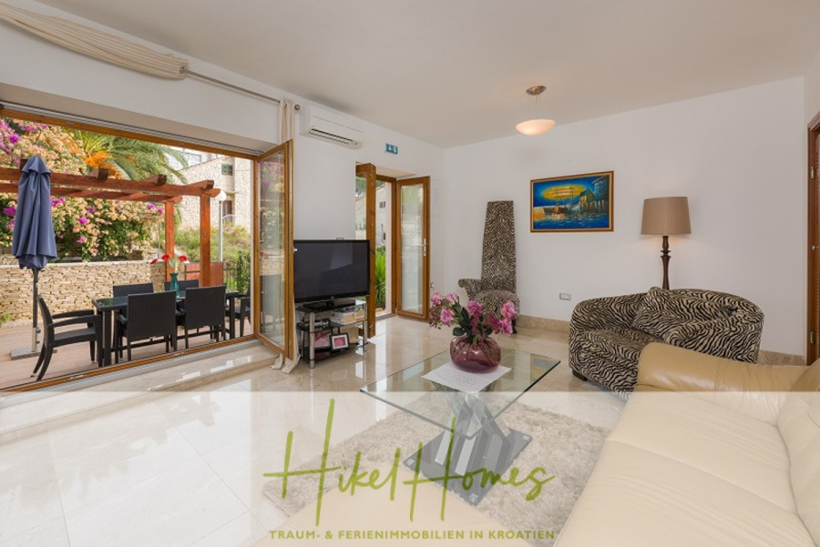 Villa mit 3 großzügigen Appartements mit jeweils 3 SZ, Küche, 2 x Pool, … - Helles Wohnzimmer mit modernen Möbeln, einem Couchtisch aus Glas, einem Fernseher und Terrassentüren, die auf eine Terrasse mit Sitzgelegenheiten im Freien und bunten Blumen führen. Das Bild zeigt das Logo von Hikel Homes am unteren Rand. (46265)