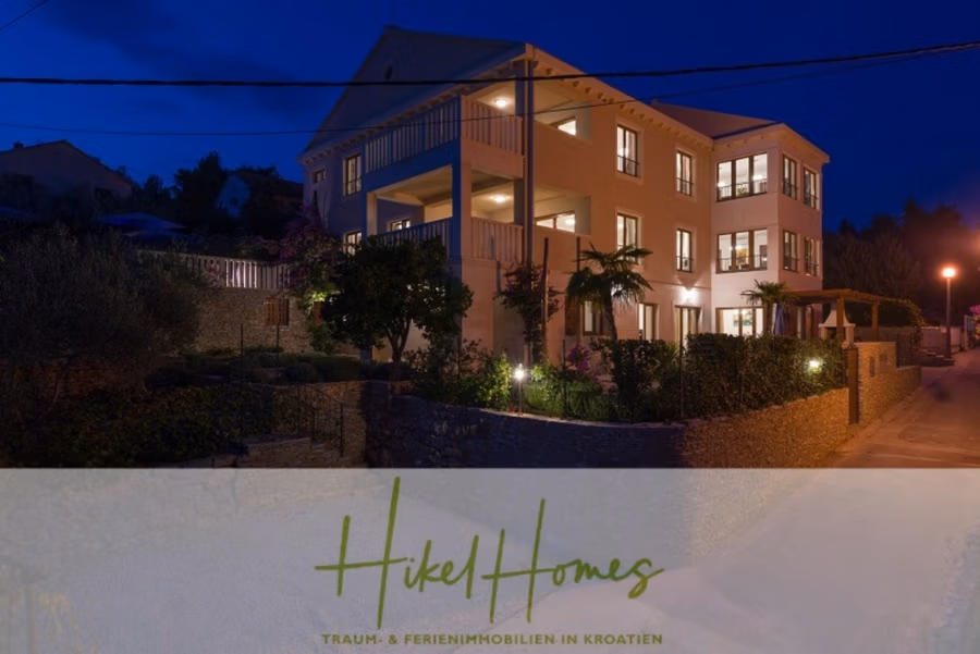 Villa mit 3 großzügigen Appartements mit jeweils 3 SZ, Küche, 2 x Pool, … - Abgebildet ist ein großes, beleuchtetes Haus mit mehreren Stockwerken und Garten bei Nacht. Das Gebäude hat eine beigefarbene Fassade und viele Fenster. Das Grundstück ist von Grünflächen umgeben. Unten befindet sich ein Logo mit der Aufschrift „Hikel Homes – TRÄUM & FERIENIMMOBILIEN IN KROATIEN“. (41130)