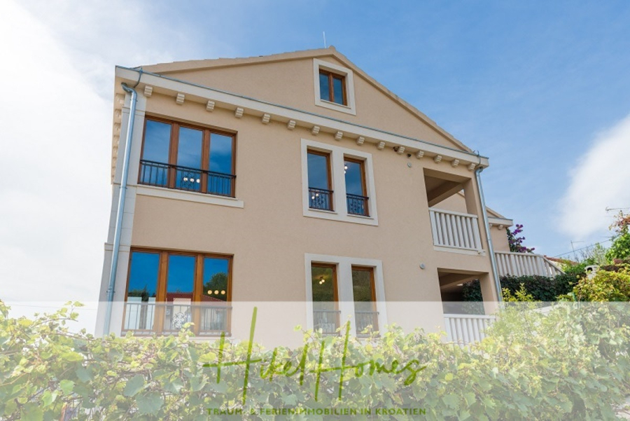 Villa mit 3 großzügigen Appartements mit jeweils 3 SZ, Küche, 2 x Pool, … - Ein beigefarbenes, dreistöckiges Haus mit großen Fenstern und Balkonen, umgeben von Grün und einem klaren blauen Himmel. Das Logo "Heil Homes" ist am unteren Rand des Bildes eingeblendet. (46262)