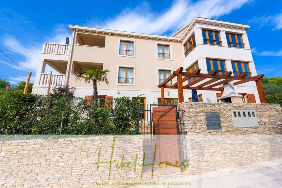 Villa mit 3 großzügigen Appartements mit jeweils 3 SZ, Küche, 2 x Pool, … - Eine moderne Villa mit beiger Fassade und mehreren Balkonen, umgeben von Grünflächen und einer Steinmauer. Am Eingang befindet sich eine Holzpergola, und über ihr wölbt sich ein blauer Himmel. (41131)